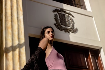 A woman wearing a pink tank top and black jacket stands confidently in front of a building entrance. The building features a large emblem above the door and tall, classical columns on one side.