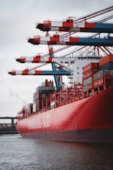 A large red container ship named HAMBURG SÜD is docked at a port with multiple cranes transferring containers. The ship is loaded with multicolored shipping containers stacked high, and the sky is overcast.