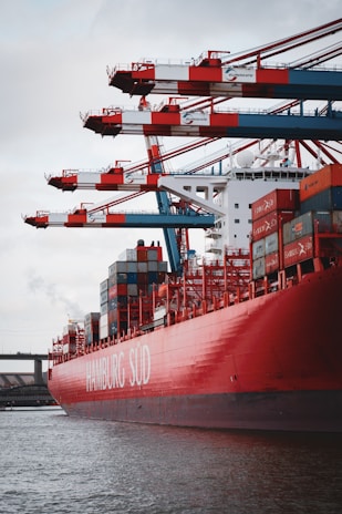 A large red container ship named HAMBURG SÜD is docked at a port with multiple cranes transferring containers. The ship is loaded with multicolored shipping containers stacked high, and the sky is overcast.
