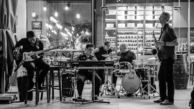 A black and white photograph captures a group of five musicians performing. The setup includes a bass player, a keyboardist, a drummer, and a saxophonist, all engaging in a lively and collaborative music session. The background features shelves stocked with wine bottles and soft ambient lighting adds to the intimate atmosphere.