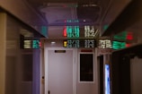 A narrow hallway with a reflective ceiling displaying digital LED signs. The signs display numeric values in green and red lights, and there is a white door marked 'WC' for a restroom. On top, a digital temperature display shows figures like 29.0°C. The ambiance is dimly lit, creating a mysterious and enclosed atmosphere.