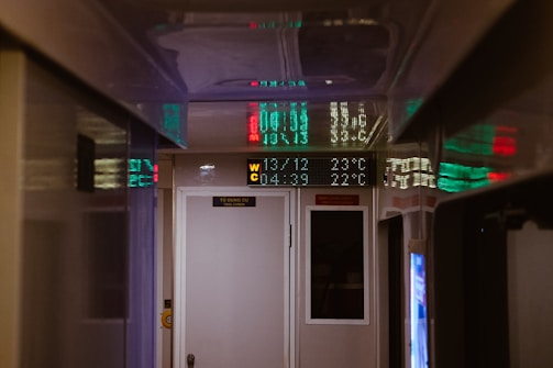 A narrow hallway with a reflective ceiling displaying digital LED signs. The signs display numeric values in green and red lights, and there is a white door marked 'WC' for a restroom. On top, a digital temperature display shows figures like 29.0°C. The ambiance is dimly lit, creating a mysterious and enclosed atmosphere.