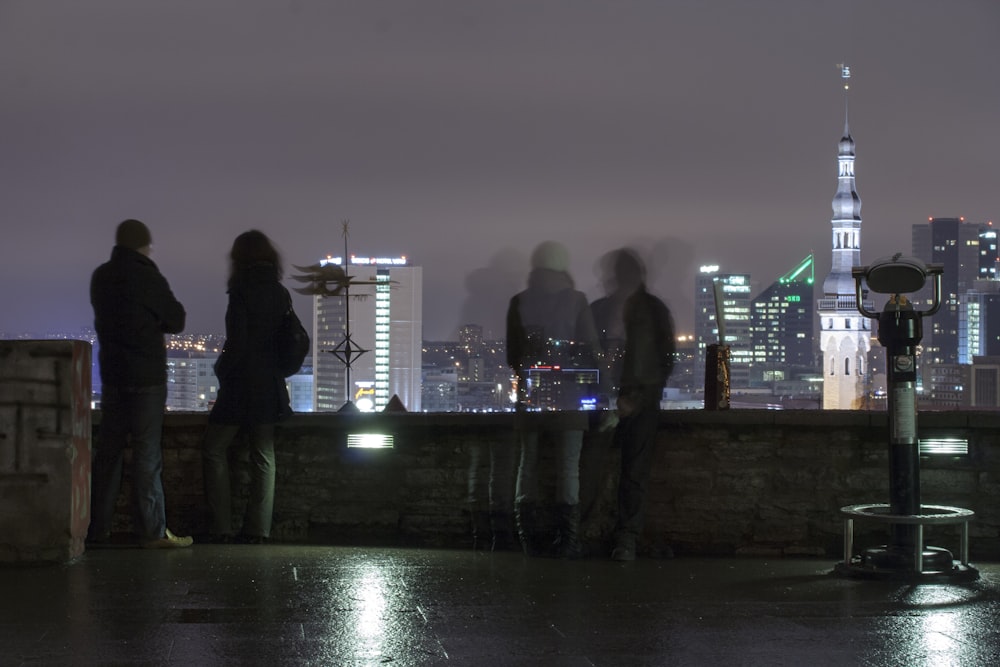 four people standing near railings viewing city with high-rise buildings during night time