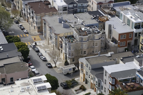 Aerial view of a residential neighborhood featuring closely packed multi-story buildings with flat and sloped roofs. Rooftop decks and balconies are visible, with several trees lining the street. A postal truck is driving down the road, and multiple parked cars are visible on the street below.