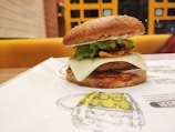 A close-up view of a gourmet burger placed on a paper mat. It features a sesame seed bun, crispy lettuce, a slice of cheese, and what appears to be a breaded patty with sauce. The background shows a brightly colored seating area with a grid-patterned wall.