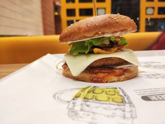 A close-up view of a gourmet burger placed on a paper mat. It features a sesame seed bun, crispy lettuce, a slice of cheese, and what appears to be a breaded patty with sauce. The background shows a brightly colored seating area with a grid-patterned wall.