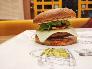 A close-up view of a gourmet burger placed on a paper mat. It features a sesame seed bun, crispy lettuce, a slice of cheese, and what appears to be a breaded patty with sauce. The background shows a brightly colored seating area with a grid-patterned wall.