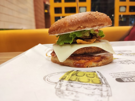A close-up view of a gourmet burger placed on a paper mat. It features a sesame seed bun, crispy lettuce, a slice of cheese, and what appears to be a breaded patty with sauce. The background shows a brightly colored seating area with a grid-patterned wall.