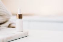 A small white dropper bottle with a golden cap sits on a marble surface. The background is softly blurred, featuring neutral tones and possibly a knitted fabric in the corner, giving the image a minimalistic and elegant appearance.