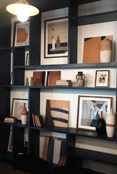 A stylish and modern shelving unit displays an assortment of framed abstract artworks, decorative vases, and neatly stacked books. The shelves are filled with a warm palette of beige, brown, and black tones, providing a sophisticated and artistic ambiance.