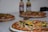 Several pizzas are arranged on wooden boards on a white surface. The pizzas are topped with various ingredients such as olives, mushrooms, bell peppers, corn, and sliced sausages. In the background, two bottles of condiments are visible, slightly out of focus.