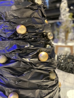 A close-up view of a stylized Christmas tree made from black material, adorned with gold and silver baubles. The tree appears to be set in a modern indoor space with additional decorative elements visible in the blurred background.