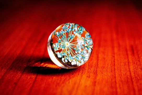 A clear spherical object, possibly a glass paperweight, containing delicate blue and white floral patterns. The sphere is placed on a warm-toned wooden surface, casting a slight shadow.