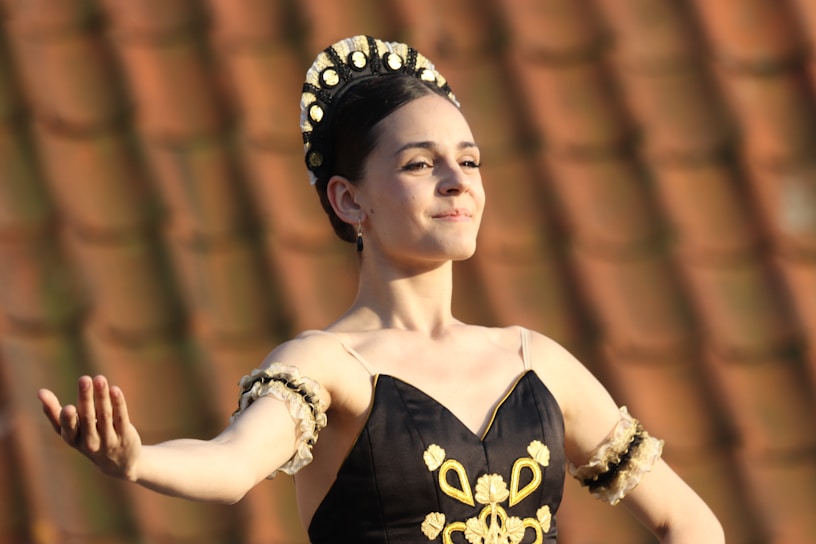 A ballet dancer dressed in traditional attire with ornate embellishments and a headpiece stands gracefully against a blurred, warm-toned background. Her arms are poised elegantly, and she appears focused and serene.