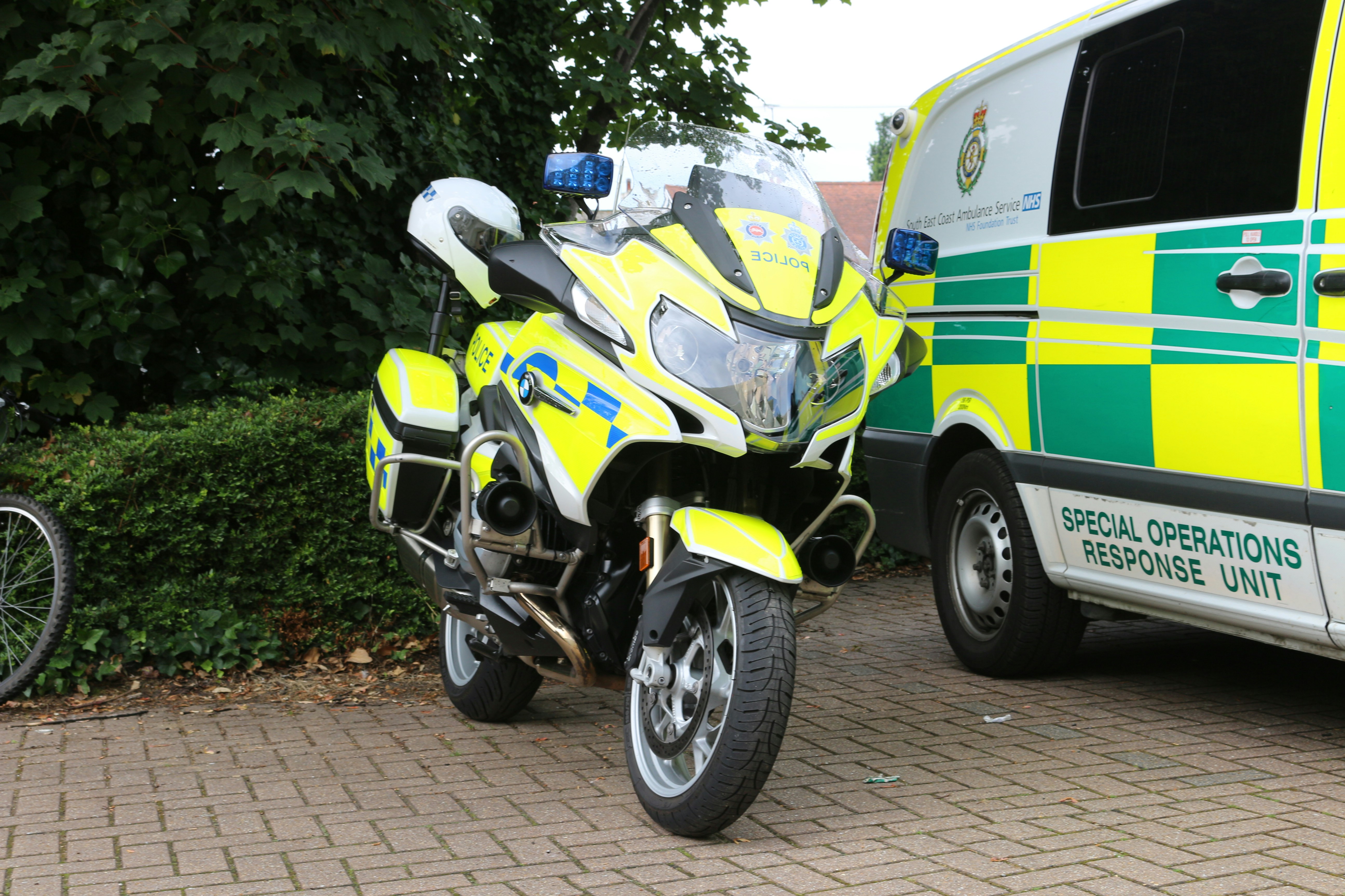 A police motorcycle with high-visibility yellow and blue markings stands parked beside a special operations response unit van. The scene is set on a paved area with greenery, including bushes and trees, in the background. A portion of a bicycle wheel is visible to the left.