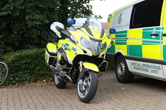 A police motorcycle with high-visibility yellow and blue markings stands parked beside a special operations response unit van. The scene is set on a paved area with greenery, including bushes and trees, in the background. A portion of a bicycle wheel is visible to the left.
