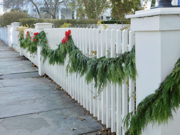 A white picket fence is decorated with green garlands and red bows, indicating a festive or holiday theme. The fence lines a stone pathway surrounded by trees and shrubs, adding a natural, serene element to the scene.