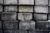 A wall constructed from large, rough-hewn stone blocks stacked in a uniform pattern. The blocks display a variety of gray tones, with some showing patches of moss and discoloration.