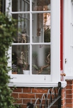 A portion of a house exterior with a window. Through the window, various plant pots and decorations can be seen. Hanging ornaments and potted plants add to the cozy and rustic atmosphere. The window frame is painted white, contrasting with the red brick wall and a small section of a red door.