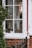A portion of a house exterior with a window. Through the window, various plant pots and decorations can be seen. Hanging ornaments and potted plants add to the cozy and rustic atmosphere. The window frame is painted white, contrasting with the red brick wall and a small section of a red door.