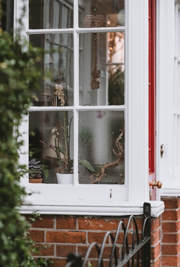 A portion of a house exterior with a window. Through the window, various plant pots and decorations can be seen. Hanging ornaments and potted plants add to the cozy and rustic atmosphere. The window frame is painted white, contrasting with the red brick wall and a small section of a red door.