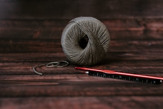 A neatly wound ball of grayish-brown yarn sits on a dark wooden surface. Next to it, a red crochet hook is placed horizontally, slightly overlapping the yarn.