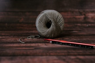 A neatly wound ball of grayish-brown yarn sits on a dark wooden surface. Next to it, a red crochet hook is placed horizontally, slightly overlapping the yarn.
