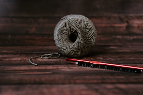 A neatly wound ball of grayish-brown yarn sits on a dark wooden surface. Next to it, a red crochet hook is placed horizontally, slightly overlapping the yarn.