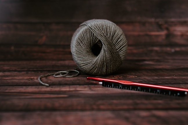 A neatly wound ball of grayish-brown yarn sits on a dark wooden surface. Next to it, a red crochet hook is placed horizontally, slightly overlapping the yarn.