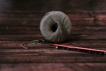 A neatly wound ball of grayish-brown yarn sits on a dark wooden surface. Next to it, a red crochet hook is placed horizontally, slightly overlapping the yarn.
