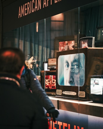 A person is standing in front of a display window that shows multiple old-fashioned televisions. On one of the screens, there is a close-up of an older man's face with the text 'Pacino' and 'NETFLIX' visible. The background includes blue curtains and another person partially visible.