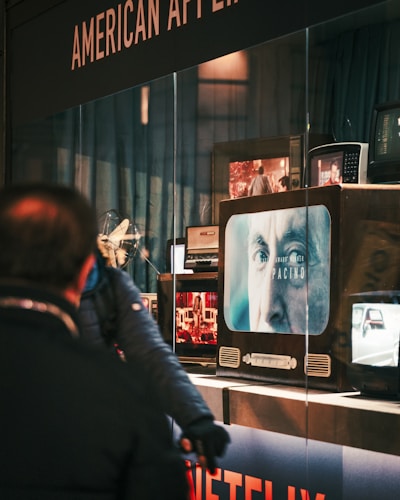 A person is standing in front of a display window that shows multiple old-fashioned televisions. On one of the screens, there is a close-up of an older man's face with the text 'Pacino' and 'NETFLIX' visible. The background includes blue curtains and another person partially visible.