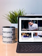 A tablet displaying a magazine website is positioned on a table, next to a mug with the word 'Bantumen' printed multiple times and a small potted plant. The tablet shows various articles and images on the screen.