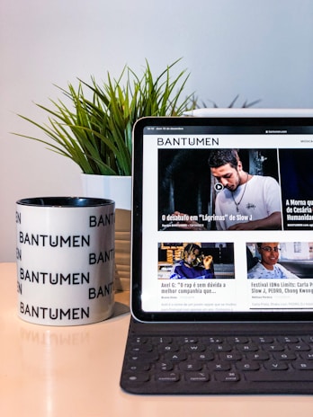 A tablet displaying a magazine website is positioned on a table, next to a mug with the word 'Bantumen' printed multiple times and a small potted plant. The tablet shows various articles and images on the screen.