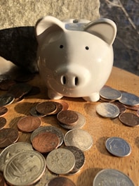 A ceramic piggy bank is surrounded by a collection of various coins on a wooden table, illuminated by warm sunlight. The piggy bank is prominently placed in the center, while the coins are scattered around, creating a sense of abundance.