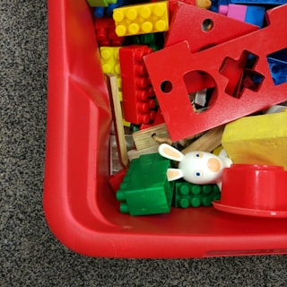 A collection of colorful building blocks, including red, yellow, green, and blue pieces, are scattered in a red plastic container. A small white toy figure with rabbit ears is visible among the blocks.