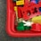 A collection of colorful building blocks, including red, yellow, green, and blue pieces, are scattered in a red plastic container. A small white toy figure with rabbit ears is visible among the blocks.