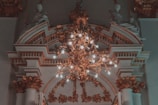 An ornate chandelier with numerous lit bulbs hangs from a detailed, decorative ceiling featuring intricate molding and architectural features. Sculpted figures sit atop the elaborate arches, adding a classical touch to the sophisticated design.