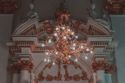 An ornate chandelier with numerous lit bulbs hangs from a detailed, decorative ceiling featuring intricate molding and architectural features. Sculpted figures sit atop the elaborate arches, adding a classical touch to the sophisticated design.