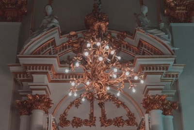 An ornate chandelier with numerous lit bulbs hangs from a detailed, decorative ceiling featuring intricate molding and architectural features. Sculpted figures sit atop the elaborate arches, adding a classical touch to the sophisticated design.