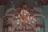 An ornate chandelier with numerous lit bulbs hangs from a detailed, decorative ceiling featuring intricate molding and architectural features. Sculpted figures sit atop the elaborate arches, adding a classical touch to the sophisticated design.