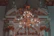 An ornate chandelier with numerous lit bulbs hangs from a detailed, decorative ceiling featuring intricate molding and architectural features. Sculpted figures sit atop the elaborate arches, adding a classical touch to the sophisticated design.