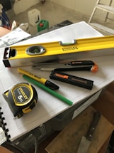 A collection of tools including a yellow level, a measuring tape, a utility knife, a green pencil, and a black marker are placed on top of some stacked papers and a plastic box. There is additional construction equipment in the background.