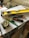 A collection of tools including a yellow level, a measuring tape, a utility knife, a green pencil, and a black marker are placed on top of some stacked papers and a plastic box. There is additional construction equipment in the background.