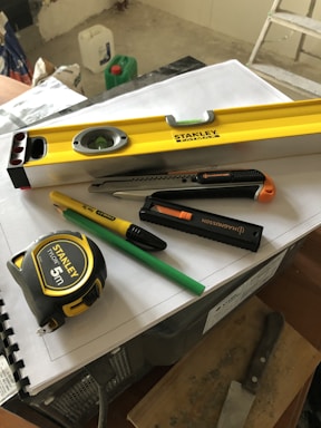 A collection of tools including a yellow level, a measuring tape, a utility knife, a green pencil, and a black marker are placed on top of some stacked papers and a plastic box. There is additional construction equipment in the background.
