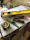 A collection of tools including a yellow level, a measuring tape, a utility knife, a green pencil, and a black marker are placed on top of some stacked papers and a plastic box. There is additional construction equipment in the background.