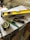 A collection of tools including a yellow level, a measuring tape, a utility knife, a green pencil, and a black marker are placed on top of some stacked papers and a plastic box. There is additional construction equipment in the background.