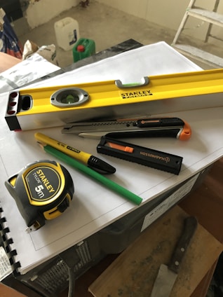 A collection of tools including a yellow level, a measuring tape, a utility knife, a green pencil, and a black marker are placed on top of some stacked papers and a plastic box. There is additional construction equipment in the background.