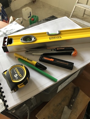 A collection of tools including a yellow level, a measuring tape, a utility knife, a green pencil, and a black marker are placed on top of some stacked papers and a plastic box. There is additional construction equipment in the background.