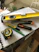 A collection of tools including a yellow level, a measuring tape, a utility knife, a green pencil, and a black marker are placed on top of some stacked papers and a plastic box. There is additional construction equipment in the background.